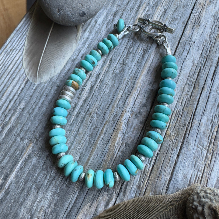 A beaded bracelet with blue Kingman turquoise rondelle beads and small Thai silver spacer beads, arranged in an oval shape on a weathered gray wood surface with decorative stones and dried leaves nearby.