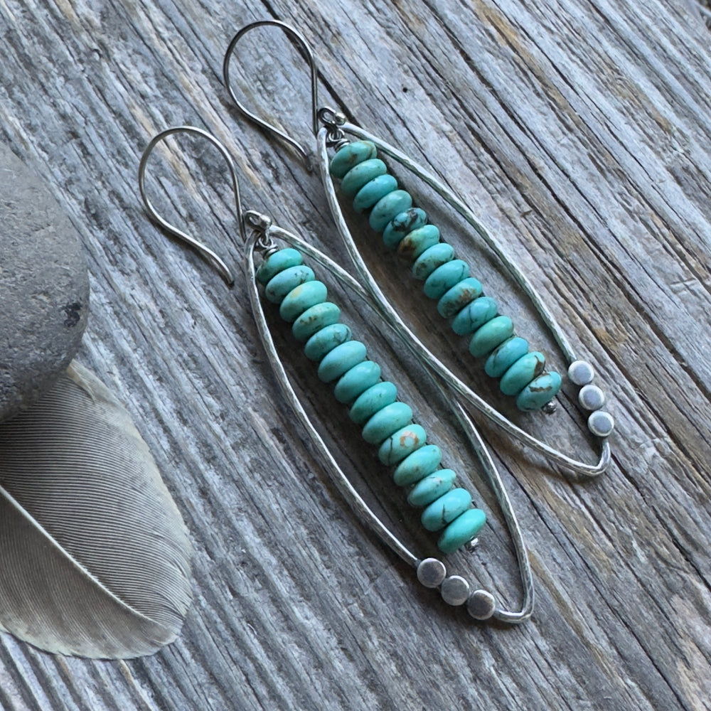 A pair of long sterling silver and Kingman turquoise earrings with open leaf and stacked turquoise rondelle design , displayed on a rustic weathered wood background with decorative feathers and a stone.