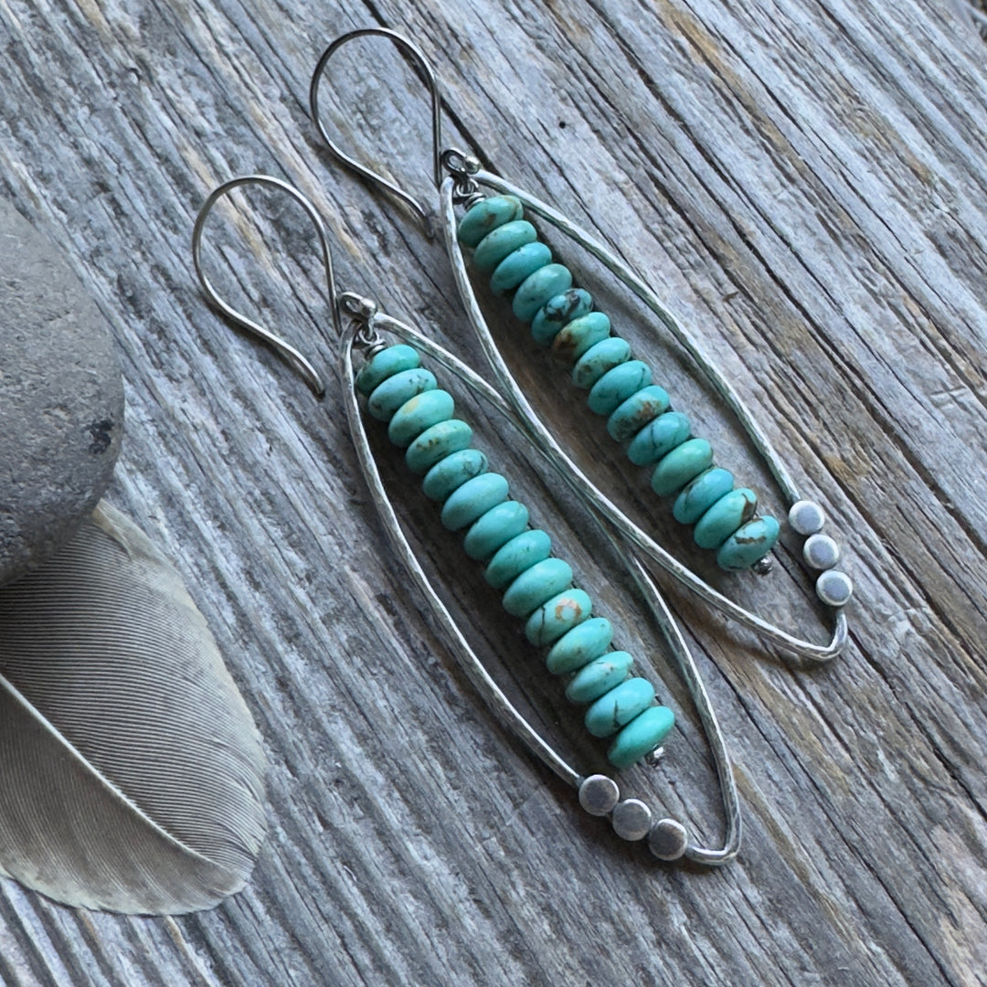 A pair of long sterling silver and Kingman turquoise earrings with open leaf and stacked turquoise rondelle design , displayed on a rustic weathered wood background with decorative feathers and a stone.