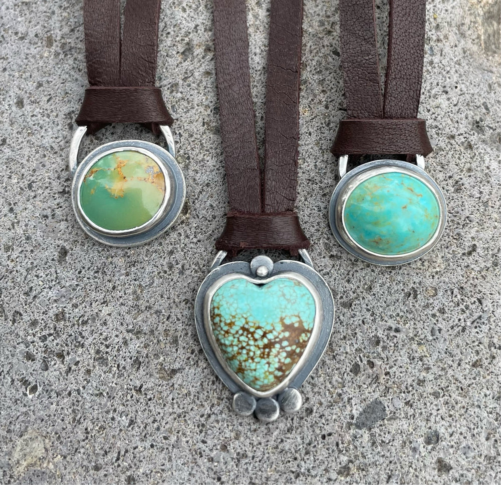 Three silver and turquoise pendant necklaces on brown leather cords, featuring two oval stones and one heart-shaped turquoise with silver accent dots, displayed on concrete.